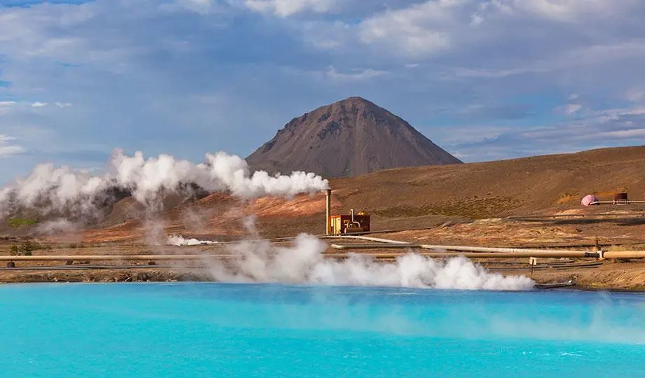 Geothermal installation with steam rising beside a turquoise pool, pipework and a volcanic hill in the background.